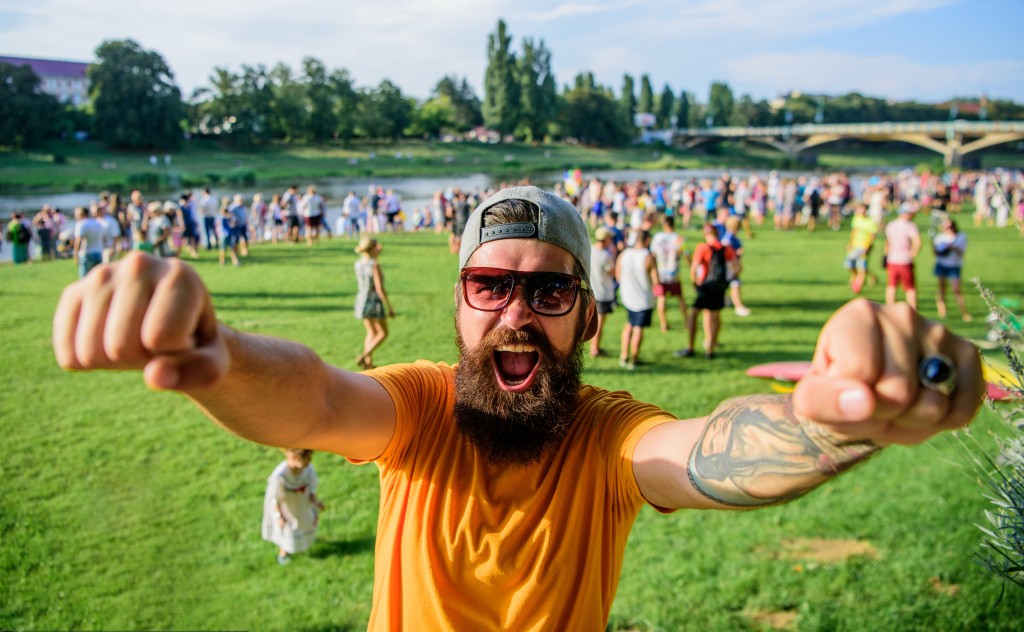Man bearded hipster in front of crowd people raise fists green riverside background. Urban event celebration. Hipster in cap happy celebrate event picnic fest or festival. Cheerful fan at summer fest.